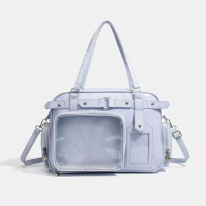 May include: A light blue handbag with a clear front pocket and a shoulder strap. The bag has a rectangular shape with a top handle and side pockets. It features silver-colored hardware and a buckle closure.