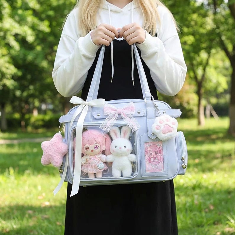 May include: A light blue ita bag with clear front pockets, decorated with pink bows and a star-shaped charm. The bag contains a doll and a white bunny plushie. A paw-shaped keychain hangs from the side.