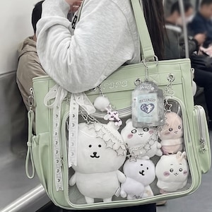 May include: A light green tote bag with a clear front window displaying several white and pink plush animal toys. The bag features a white lace ribbon, a silver star-shaped chain, and a rectangular card holder with the text I LOVE YOU.