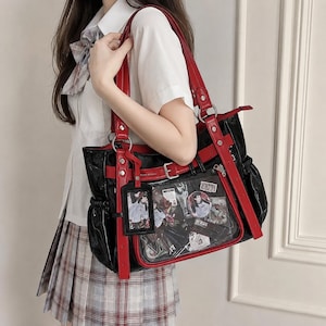May include: A black tote bag with red trim and straps, featuring a clear front pocket filled with various anime-style character cards and circular pins. The bag includes a matching black and red luggage-style tag hanging from a silver ring.