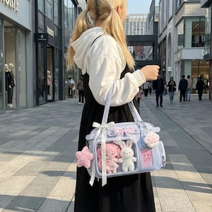 May include: A light blue handbag with a clear front panel, featuring a pink doll, a white bunny plush, and a pink star-shaped charm. The bag has a bow and a paw-shaped charm. The person is wearing a black dress and a white hoodie.