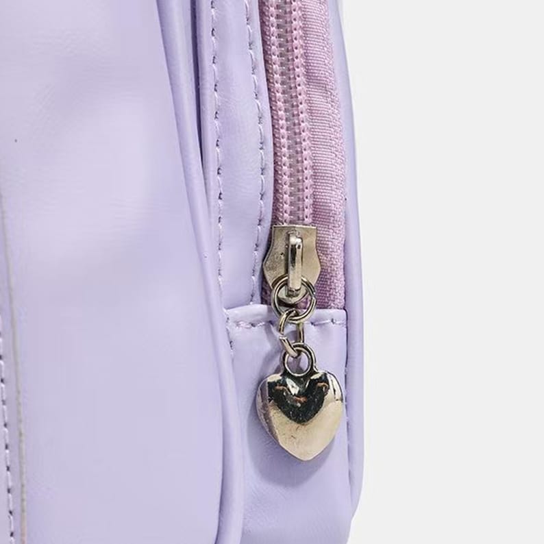 May include: Close-up of a lavender-colored bag with a matching zipper and a silver heart-shaped charm. The zipper is a lighter shade of purple, and the bag appears to be made of a smooth, synthetic material.