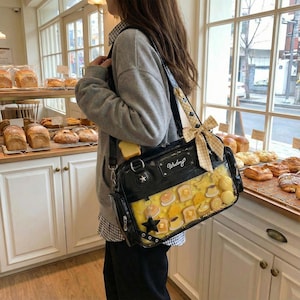 May include: Black shoulder bag with a clear front panel filled with miniature food items like pancakes and toast. The bag has a star-shaped charm and a bow. The word "Wudong" is printed on the bag.