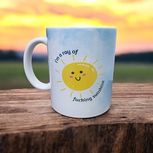 May include: White ceramic mug with a blue and white watercolor background. The mug features a yellow smiling sun with the text "I'm a ray of fucking sunshine".
