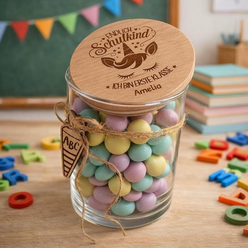 Pode incluir: Um frasco de vidro transparente cheio de doces em tons pastel, amarrado com barbante e coberto com uma tampa de madeira. A tampa &eacute; gravada com um desenho de unic&oacute;rnio e as palavras "Endlich Schulkind" e "Ich bin erste Klasse Amelia."