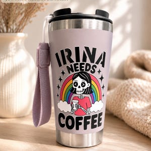 Könnte beinhalten: Ein 500 ml Thermobecher aus Edelstahl mit schwarzem Deckel und lavendelfarbener Außenseite. Der Becher zeigt eine Grafik eines Skeletts mit Kaffeetasse vor einem Regenbogen und Wolken, den Text IRINA NEEDS COFFEE und eine passende Trageschlaufe.
