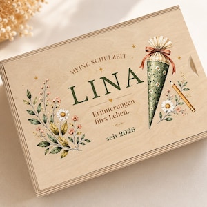 Könnte beinhalten: Eine rechteckige Holzkiste mit dem aufgedruckten Text MEINE SCHULZEIT, LINA, Erinnerungen fürs Leben und seit 2026. Das Design zeigt eine grüne Schultüte, einen Bleistift sowie florale Illustrationen mit Gänseblümchen.