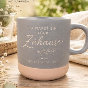 Könnte beinhalten: Ein zweifarbiger Keramikbecher mit einem gesprenkelten grauen oberen Teil und einem hellpfirsichfarbenen Boden. Der Becher trägt die Gravur "DU WARST EIN STÜCK Zuhause" über einer Blattillustration und "Spatzengruppe · 2026".