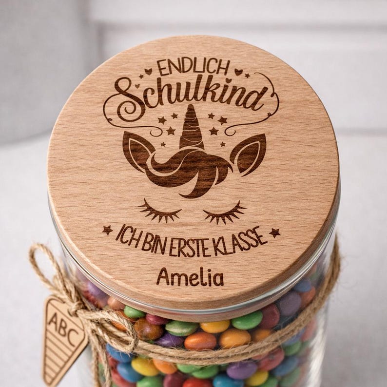 Pode incluir: Um frasco de vidro cheio de doces coloridos, coberto com uma tampa de madeira. A tampa &eacute; gravada com um desenho de unic&oacute;rnio e as palavras "ENDLICH Schulkina" e "ICH BIN ERSTE KLASSE". O frasco &eacute; amarrado com barbante e tem uma pequena etiqueta de madeira.