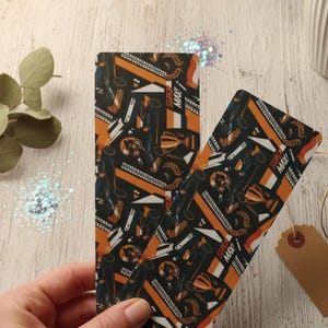 Può includere: Due segnalibri rettangolari con un motivo nero e arancione con illustrazioni a tema racing e la scritta "SUPER MAX!". I segnalibri sono tenuti su una superficie di legno chiaro.