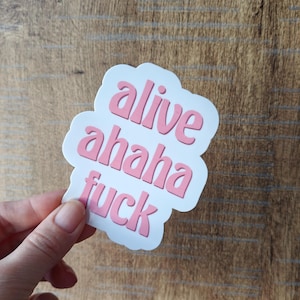Puede incluir: Una pegatina blanca con un borde en forma de nube. La pegatina presenta las palabras "alive ahaha fuck" en rosa, con letras estilizadas. La pegatina se sostiene sobre un fondo de madera.