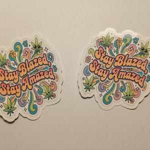 May include: Two stickers with a colorful design featuring the text "Stay Blazed, Stay Amazed". The stickers have a white border and include cartoon cannabis leaves, flowers, and swirling lines in pastel colors.