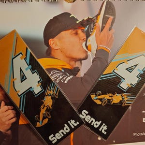 Può includere: Due segnalibri rettangolari arancioni e neri con il numero 4 e una grafica di auto da corsa. Il testo "Send it." è stampato in basso. Lo sfondo presenta una persona che bacia una scarpa.