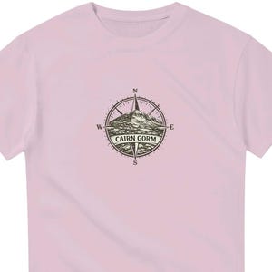 May include: A light pink short-sleeve t-shirt featuring a dark brown graphic on the chest. The graphic depicts a compass rose design with a mountain illustration in the center and the text CAIRN GORM printed across the middle of the compass.