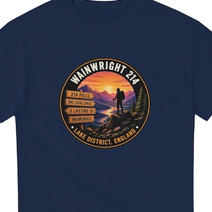 May include: A navy blue t-shirt featuring a circular graphic of a hiker standing on a rocky cliff overlooking a mountain valley at sunset. The text reads WAINWRIGHT 214, 214 FELLS, ONE CHALLENGE, A LIFETIME OF MEMORIES, and LAKE DISTRICT, ENGLAND.