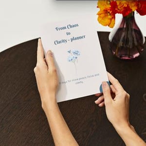 Op de afbeelding: Een witte planner met de tekst From Chaos to Clarity - planner in donkerblauw gedrukt, voorzien van een kleine illustratie van lichtblauwe bloemen en de tekst 6 days for more peace, focus and clarity onderaan met een blauw cirkelaccent.