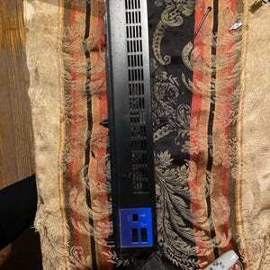 Peut inclure: Un composant électronique noir avec une série d'ouvertures verticales et un panneau bleu avec deux ouvertures rectangulaires. Un marqueur permanent Sharpie blanc est à côté du composant.