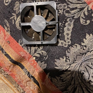 Peut inclure: Un petit ventilateur de forme carrée pour ordinateur, avec un cadre argenté et des pales marron. Le ventilateur possède un moyeu central et est conçu pour refroidir les composants électroniques. Il repose sur un tissu à motifs avec des accents rouges, oranges et dorés.