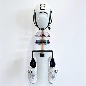 Può includere: Un supporto a parete con un montante verticale in legno, un casco bianco in cima, due paia di occhiali da ciclismo e una mensola in metallo nero che sostiene un paio di scarpe da ciclismo bianche con chiusure a rotella e dettagli in rete.