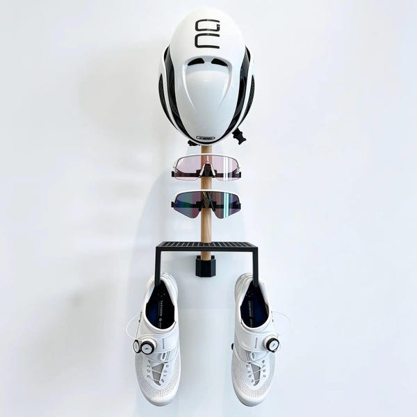 May include: A wall-mounted storage rack featuring a vertical wooden post with a white helmet at the top, two pairs of cycling sunglasses, and a black metal shelf holding a pair of white cycling shoes with dial closures and mesh detailing.