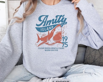 May include: A light gray sweatshirt with a graphic featuring a shark with an open mouth, the words "Amity Island" in blue, and the year "1975". Below the shark are the words "Clear Skies+Gentle Surf Warm Water". The sweatshirt is labeled "Sport Gray".
