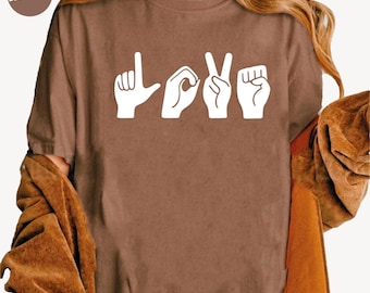 May include: Espresso-colored t-shirt with the word "LOVE" spelled out in white hand gestures. The shirt is a solid color and has short sleeves. A brown jacket is draped over the arm.