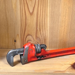 May include: A heavy-duty, adjustable pipe wrench with a bright red handle and a black jaw. The wrench is lying on a wooden surface, with the text "HEAVY DUTY" visible on the handle.