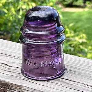 May include: A vintage, purple glass insulator with a rounded top and a flared base. The insulator has a glossy finish and the text "W.C. MCO." is embossed on the side. The object is sitting on a weathered wooden surface.