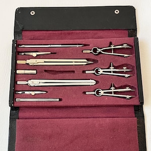 May include: A vintage drafting set in a black leatherette case. The set includes several silver-toned compasses, dividers, and other drawing tools, nestled in a burgundy velvet-lined interior. The case is open, revealing the tools neatly arranged.