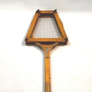 Puede incluir: Una raqueta de tenis antigua de madera con una cabeza rectangular y una red de malla. El mango es largo y delgado, con un agarre envuelto cerca de la base. La madera tiene un tono cálido y natural, y la raqueta muestra signos de edad.