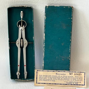 Può includere: Compasso in metallo vintage con accenti rossi, aperto in una scatola verde acqua. La scatola è aperta, rivelando lo strumento e una scheda di garanzia. La scheda recita "Garanzia" e "Punti divisori".