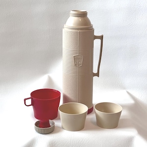 Puede incluir: Un termo beige vintage de la marca Thermos con asa, acompañado de una taza roja, dos tazas beige y una tapa de taza. El termo tiene una superficie texturizada y el logotipo de Thermos.