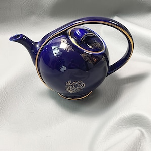 May include: A navy blue ceramic teapot with a gold trim and floral design. The teapot has a curved handle and a lid with a small knob. The gold accents highlight the elegant shape of the teapot.
