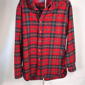 May include: A red, green, and navy plaid button-down shirt. The long-sleeved shirt has a button-down collar and a red lining inside the collar. The shirt is displayed on a hanger against a white background.
