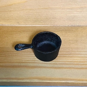 Puede incluir: Una pequeña taza medidora redonda de hierro fundido negro con un mango corto y resistente que presenta un orificio circular en el extremo. El interior de la taza tiene un acabado mate texturizado, mientras que el exterior es liso y negro.
