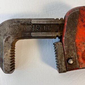 May include: Close-up of a vintage pipe wrench. The wrench is primarily metal with a rusty, worn appearance. The handle is painted bright orange. The jaws are open, displaying size markings: 1/2, 3/4, and 1 1/2.