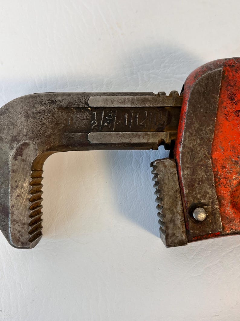 May include: Close-up of a vintage pipe wrench. The wrench is primarily metal with a rusty, worn appearance. The handle is painted bright orange. The jaws are open, displaying size markings: 1/2, 3/4, and 1 1/2.