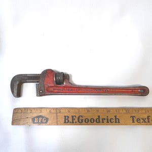 May include: A red Ridgid pipe wrench with a black jaw and adjustable nut. The wrench is marked "Ridgid" and "14". A wooden ruler with "B.F.Goodrich" printed on it is below the wrench.