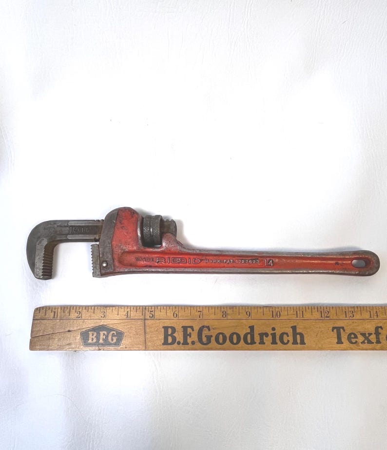May include: A red Ridgid pipe wrench with a black jaw and adjustable nut. The wrench is marked "Ridgid" and "14". A wooden ruler with "B.F.Goodrich" printed on it is below the wrench.