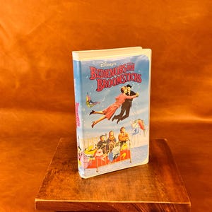 May include: A Disney's Bedknobs and Broomsticks VHS tape featuring a light blue cover with an illustration of two people flying above a brass bed frame containing three children and various animated sea creatures against a white background.
