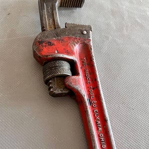 May include: A vintage red and silver pipe wrench. The wrench has a textured handle and adjustable jaws. The text "THE RIDGE TOOL CO. ELYRIA, OHIO U.S.A." is printed on the handle. The wrench is a tool for plumbing and mechanical work.