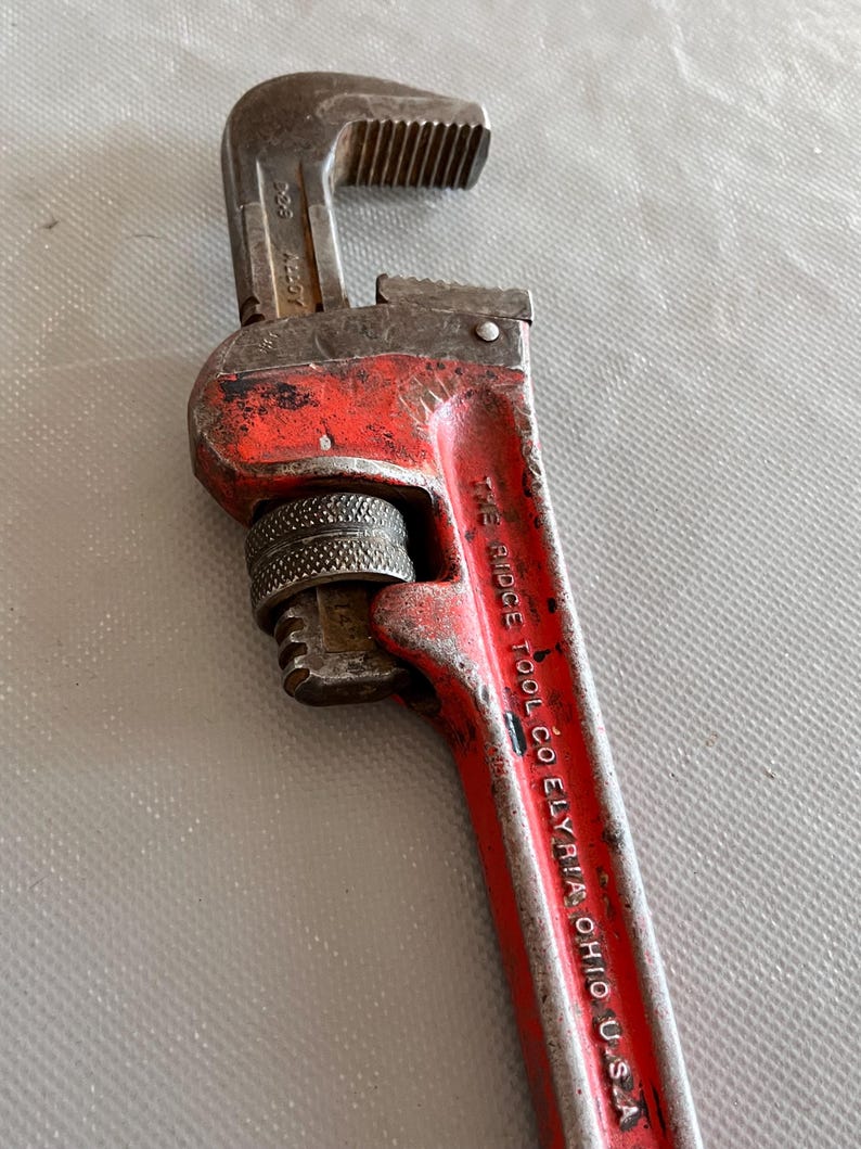 May include: A vintage red and silver pipe wrench. The wrench has a textured handle and adjustable jaws. The text "THE RIDGE TOOL CO. ELYRIA, OHIO U.S.A." is printed on the handle. The wrench is a tool for plumbing and mechanical work.