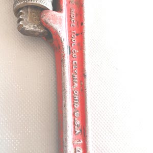 May include: A vintage red pipe wrench with the text "THE RIDGE TOOL CO ELYRIA OHIO U.S.A. 14" printed on the handle. The wrench has a silver adjustable jaw and a worn appearance, suggesting it's been used.