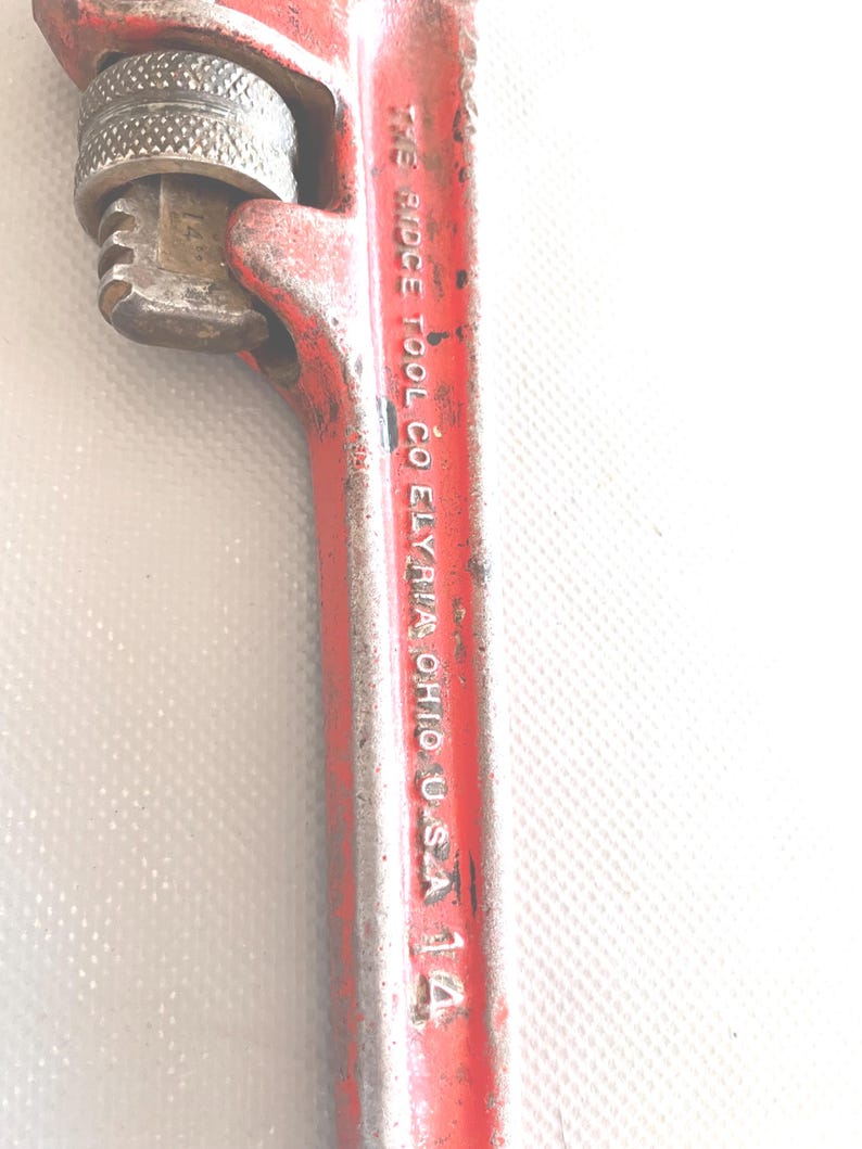 May include: A vintage red pipe wrench with the text "THE RIDGE TOOL CO ELYRIA OHIO U.S.A. 14" printed on the handle. The wrench has a silver adjustable jaw and a worn appearance, suggesting it's been used.