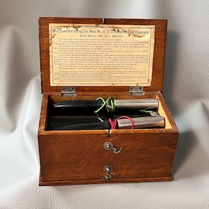 Puede incluir: Una antigua caja de aparatos médicos de madera con tapa con bisagras, que contiene instrumentos cilíndricos negros y plateados. La caja tiene una hoja de instrucciones impresa dentro de la tapa y dos pequeños cajones con cerraduras de ojo de cerradura. El texto de la tapa dice "Directions for Using the New No. 4. D. D. Home Medical Apparatus".