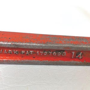 May include: Close-up of a weathered, red metal tool. The tool has the text "MARK PAT 1727623 14" embossed on its surface. The metal shows signs of age and wear.
