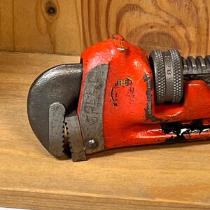 May include: Close-up of a vintage red pipe wrench. The wrench has a black jaw and a textured handle. The tool is resting on a wooden surface, showcasing its well-worn condition and classic design.