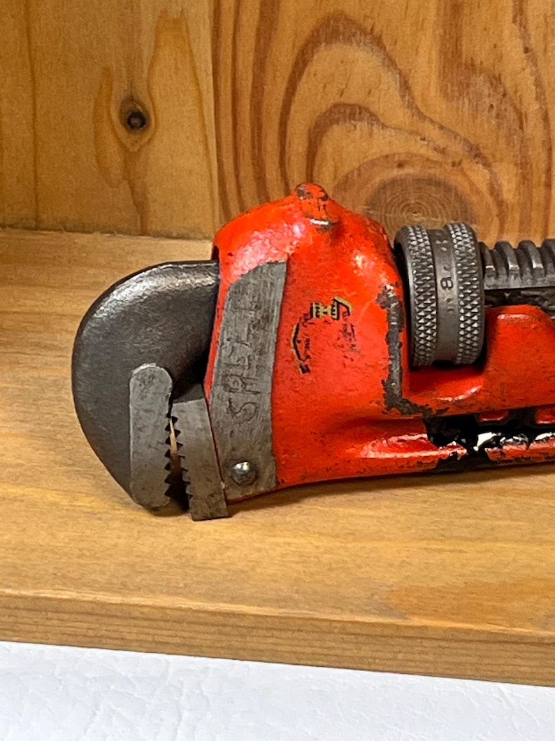 May include: Close-up of a vintage red pipe wrench. The wrench has a black jaw and a textured handle. The tool is resting on a wooden surface, showcasing its well-worn condition and classic design.