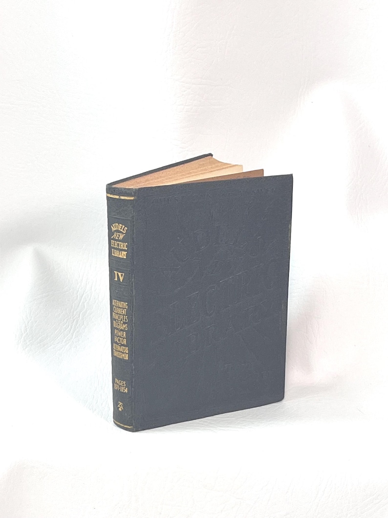 Puede incluir: Un libro antiguo con una cubierta azul oscuro, titulado "Models New Electric Library IV". El lomo del libro muestra texto adicional sobre los principios de la corriente alterna y el factor de potencia. Las p&aacute;ginas est&aacute;n envejecidas y el libro est&aacute; sobre un fondo blanco.