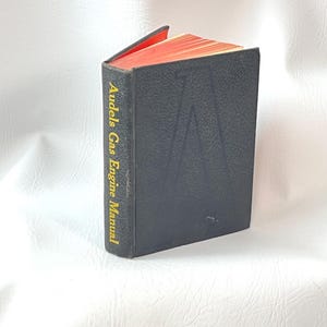 May include: A black hardcover book titled "Audels Gas Engine Manual" with gold lettering. The pages have red edges. The book is closed, with a small portion of the red pages visible. The cover has a textured appearance.